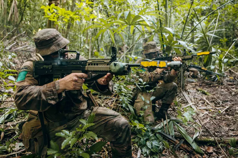 Two soldiers in camouflage gear aiming rifles while crouched in dense jungle foliage during a tactical exercise.