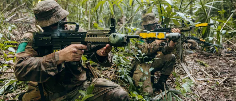 Two soldiers in camouflage gear aiming rifles while crouched in dense jungle foliage during a tactical exercise.