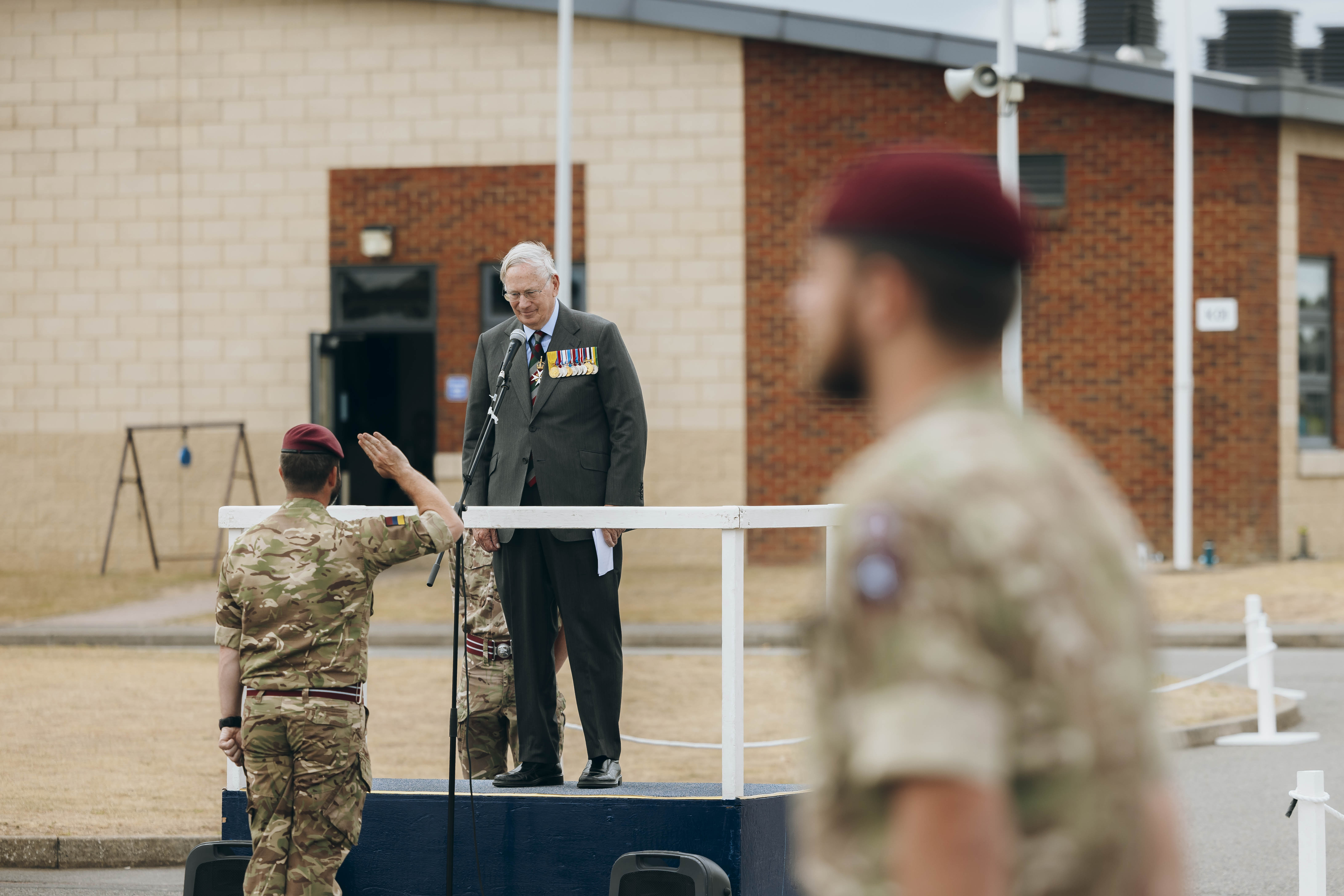 His Royal Highness the Duke of Gloucester stands wearing a suit. In front of him, a soldier wearing camouflage uniform and a maroon berets salutes.