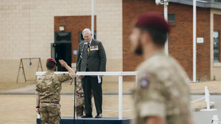 His Royal Highness the Duke of Gloucester stands wearing a suit. In front of him, a soldier wearing camouflage uniform and a maroon berets salutes.