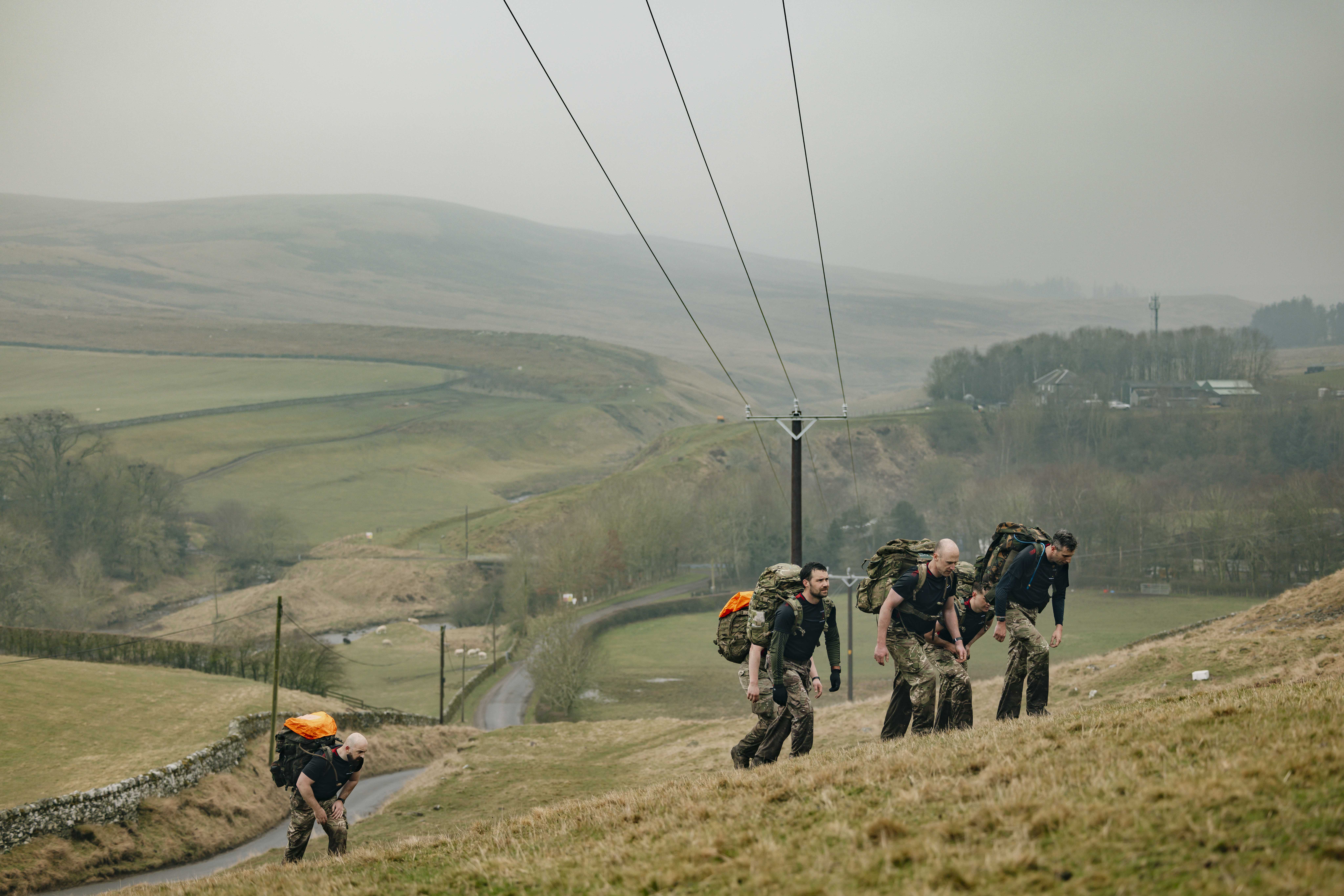 Soldier are seen carrying their gear in large green camouflage backpacks walking up a hill in the Otterburn countryside in the wet weather. 