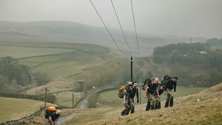 Soldier are seen carrying their gear in large green camouflage backpacks walking up a hill in the Otterburn countryside in the wet weather.