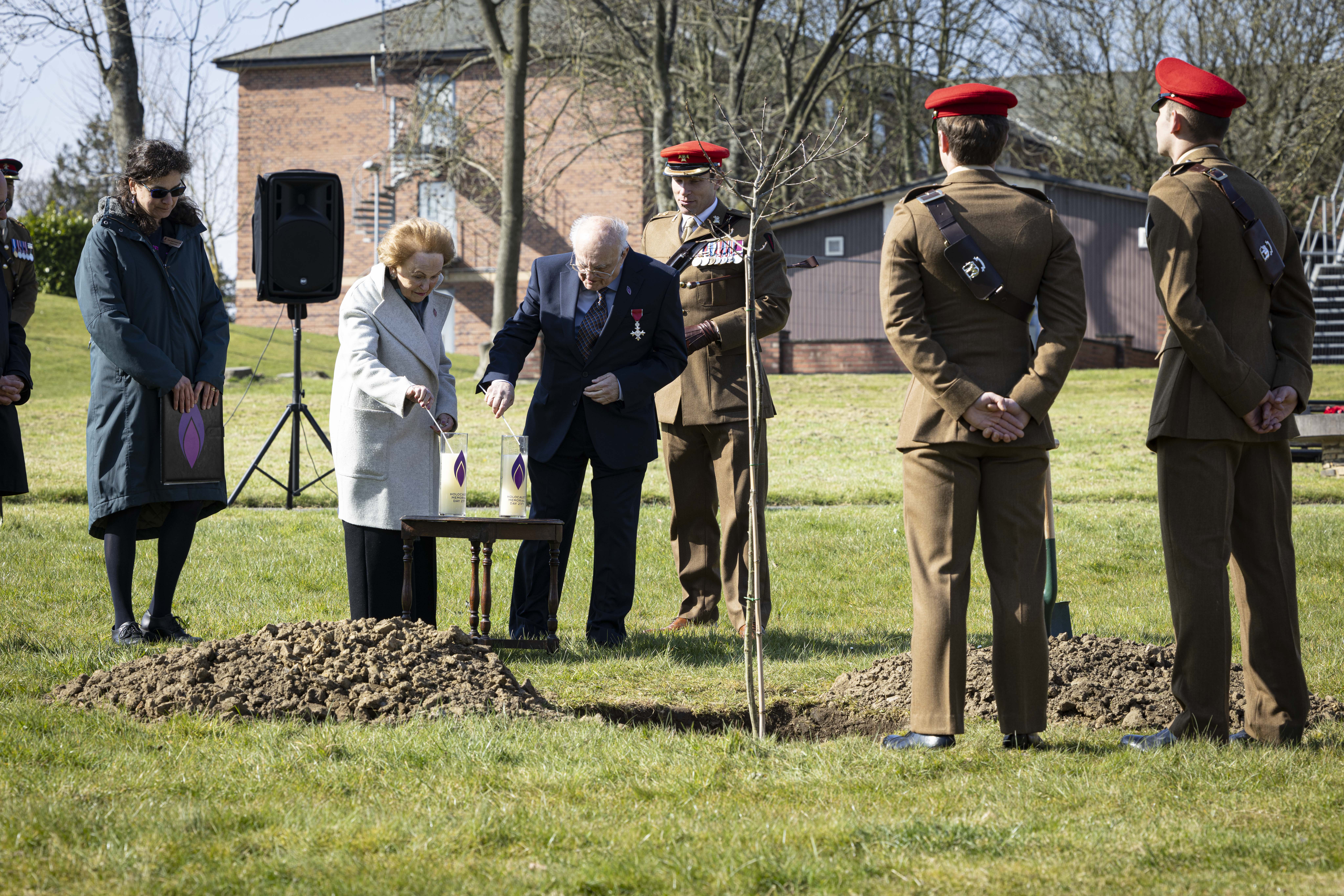 Holocaust survivors Mala Tribich MBE and Dr Alfred Garwood MBE light Holocaust candles on an army camp during a memorial service. Surrounded by several soldiers in their number 2 uniforms.  