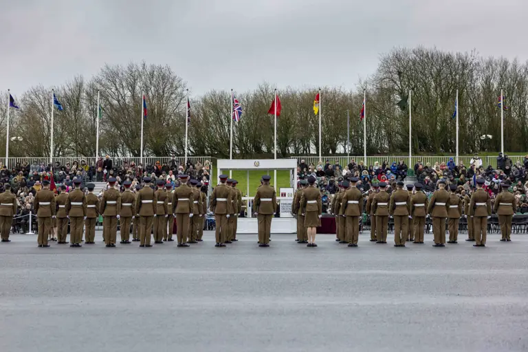 Soldiers in brown uniforms stand in formation before a seated crowd. Flags wave above, and trees are in the background under a grey sky.