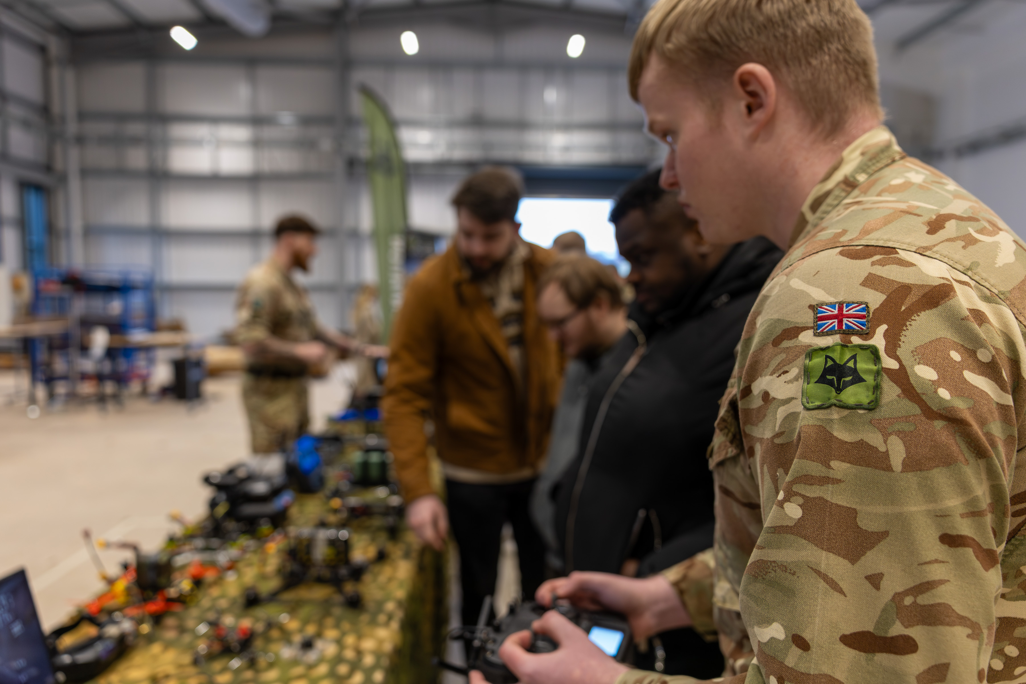 British soldier in camouflage uniform demonstrating drone controls to a group inside a large industrial space.