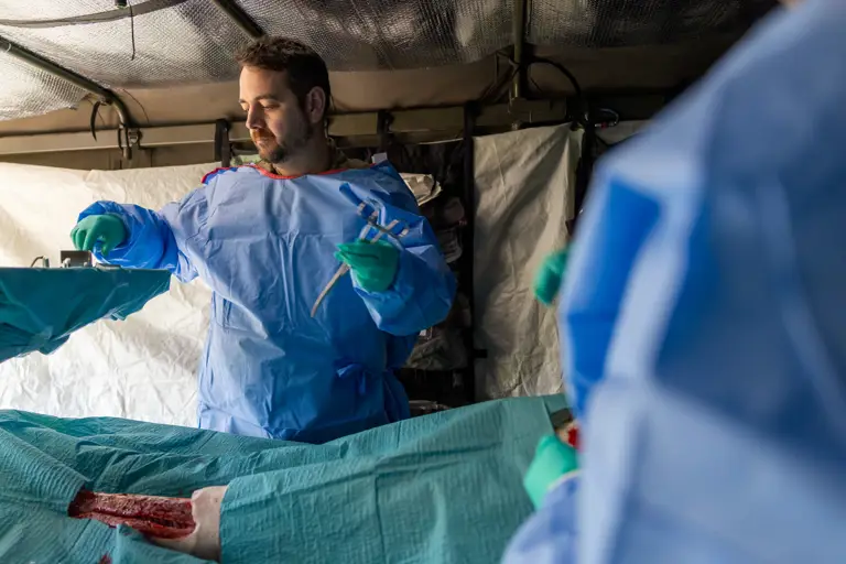 Medical personnel in protective gear preparing to treat a severe leg injury inside a field hospital tent.