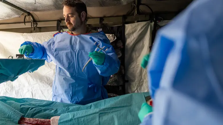 Medical personnel in protective gear preparing to treat a severe leg injury inside a field hospital tent.
