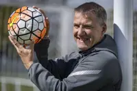A person stands beside a football goal post, smiling while holding a football. The net and field are visible.