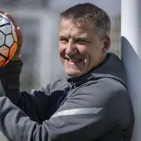 A person stands beside a football goal post, smiling while holding a football. The net and field are visible.