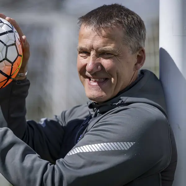 A person stands beside a football goal post, smiling while holding a football. The net and field are visible.