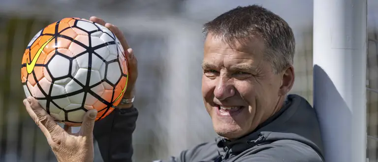A person stands beside a football goal post, smiling while holding a football. The net and field are visible.