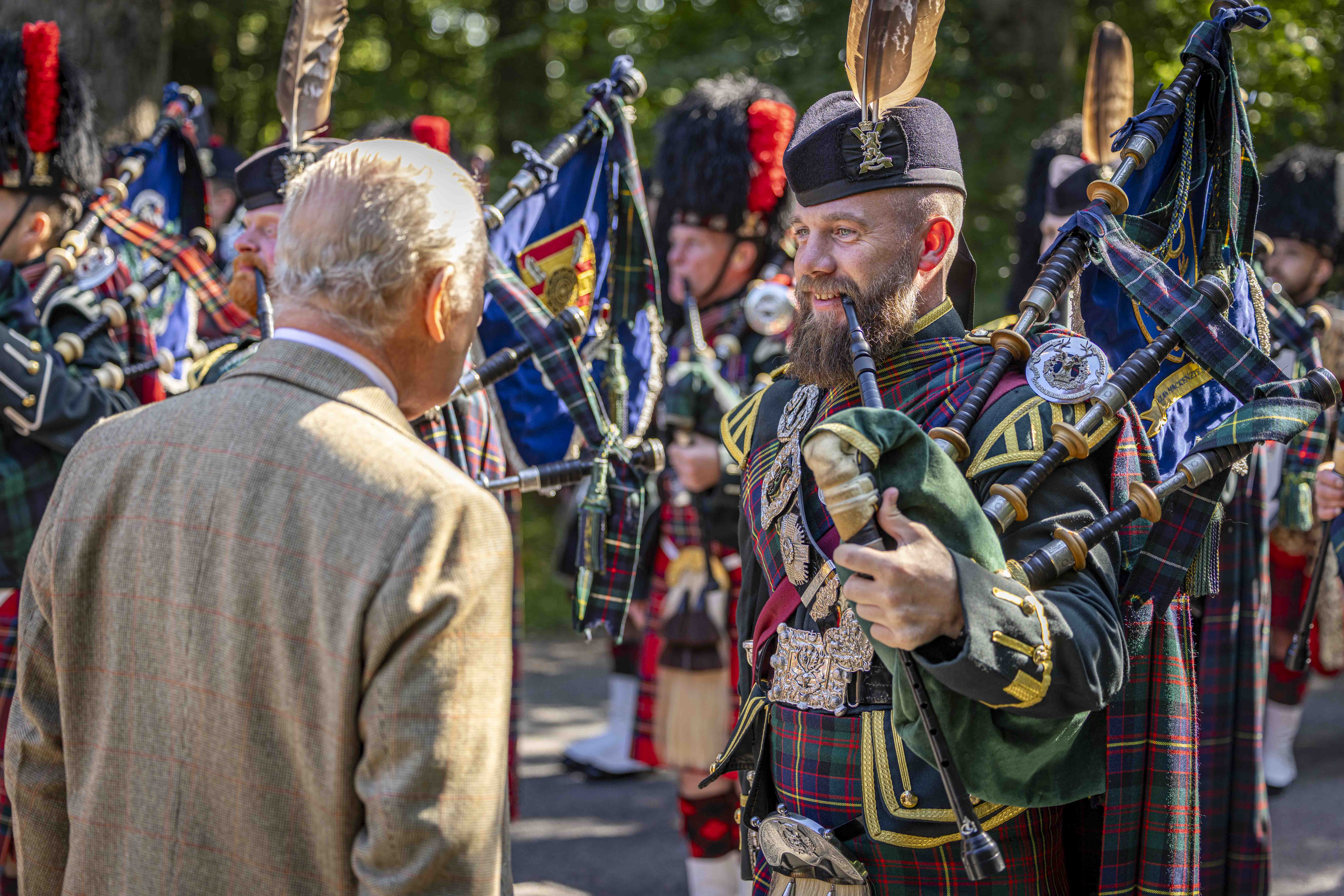 His Majesty The King in a tweed jacket observes a group of bagpipers dressed in traditional Scottish attire outdoors.