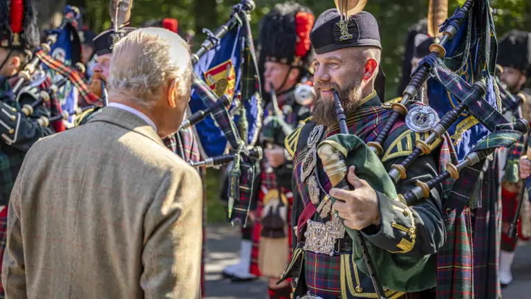 His Majesty The King in a tweed jacket observes a group of bagpipers dressed in traditional Scottish attire outdoors.