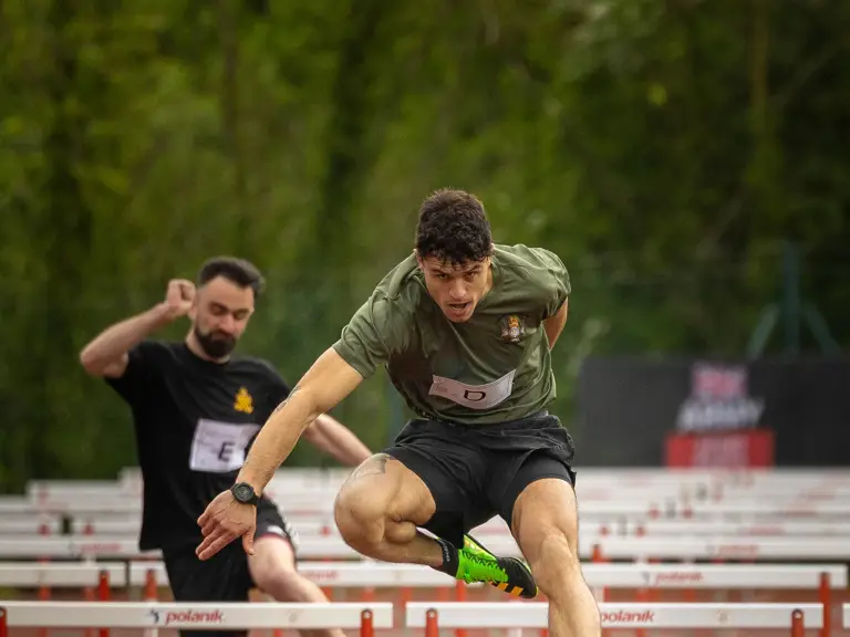 Two people are competing in a track and field hurdle race. The focus is on one participant in the foreground.