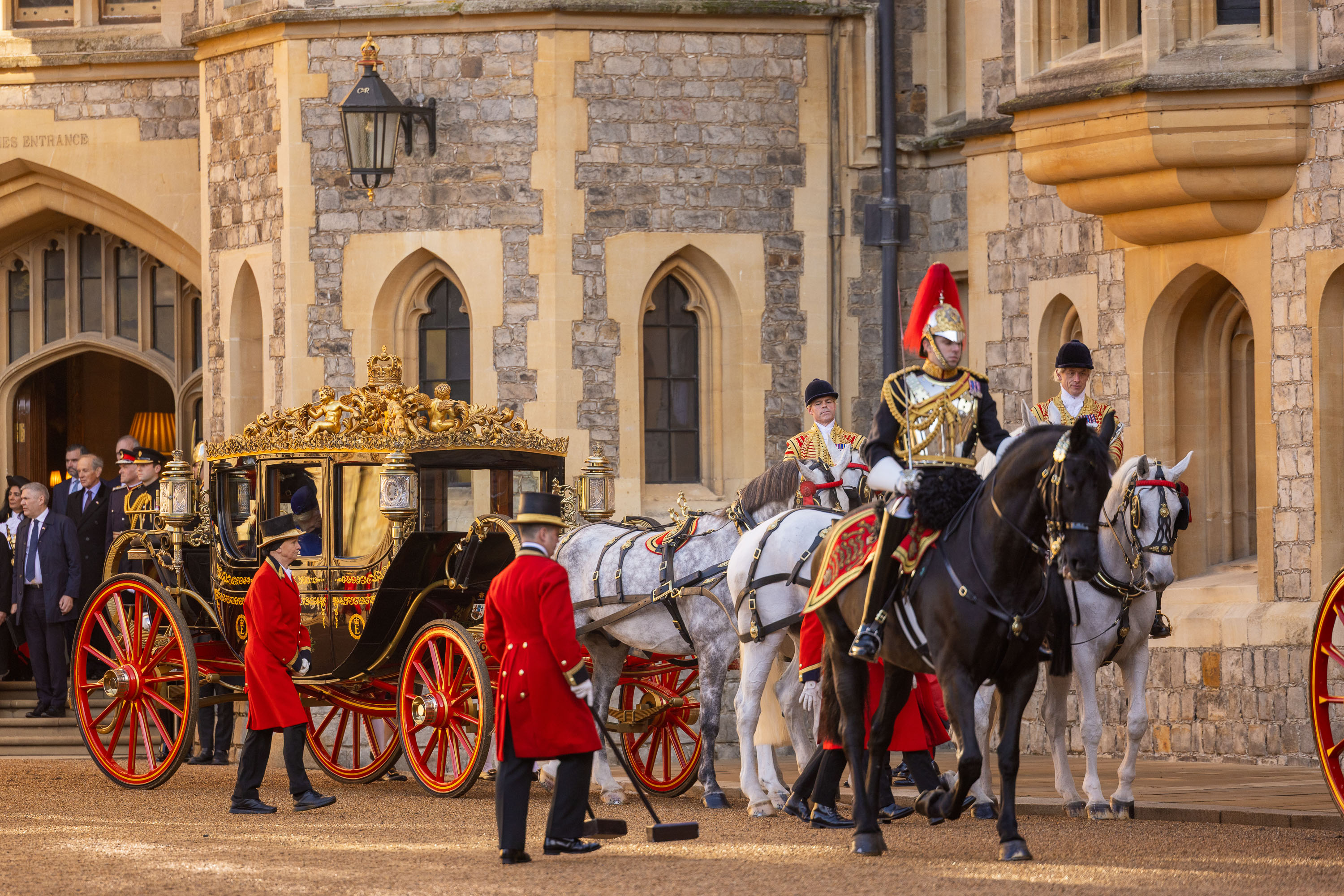 Ornate carriage with golden details, pulled by four white horses, escorted by guards in red uniforms and a knight. Historic castle backdrop. Regal ambiance.