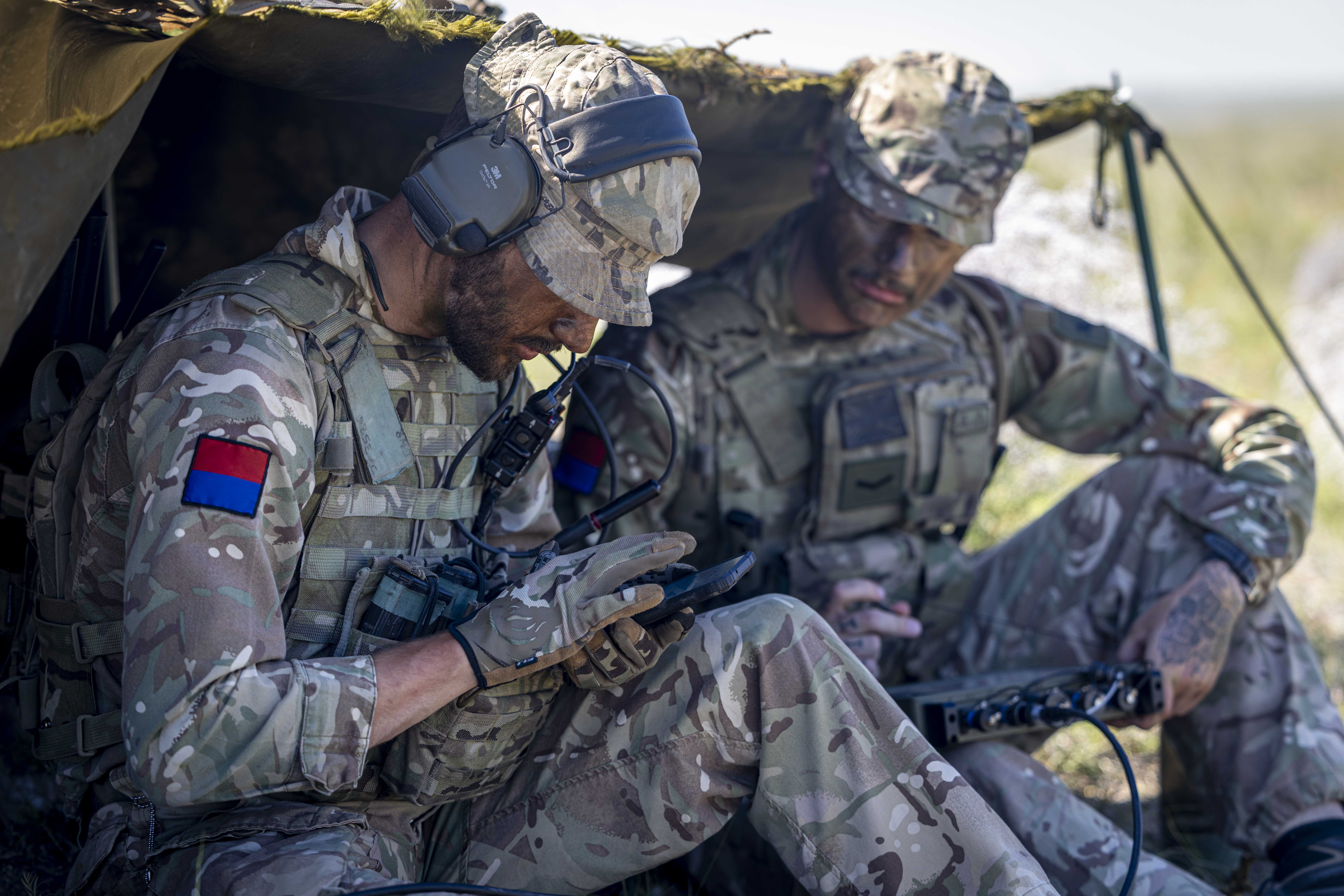 Two soldiers in camouflage uniforms sit under a makeshift shelter, using communication equipment.
