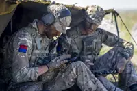 Two soldiers in camouflage uniforms sit under a makeshift shelter, using communication equipment.
