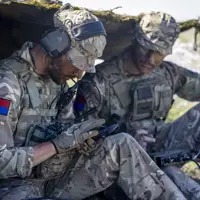Two soldiers in camouflage uniforms sit under a makeshift shelter, using communication equipment.