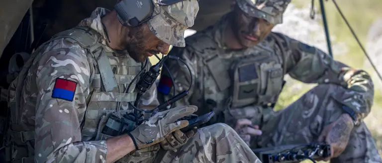 Two soldiers in camouflage uniforms sit under a makeshift shelter, using communication equipment.