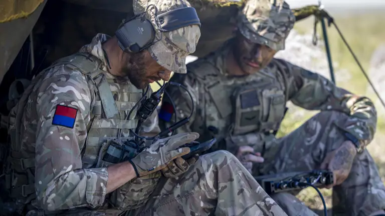 Two soldiers in camouflage uniforms sit under a makeshift shelter, using communication equipment.