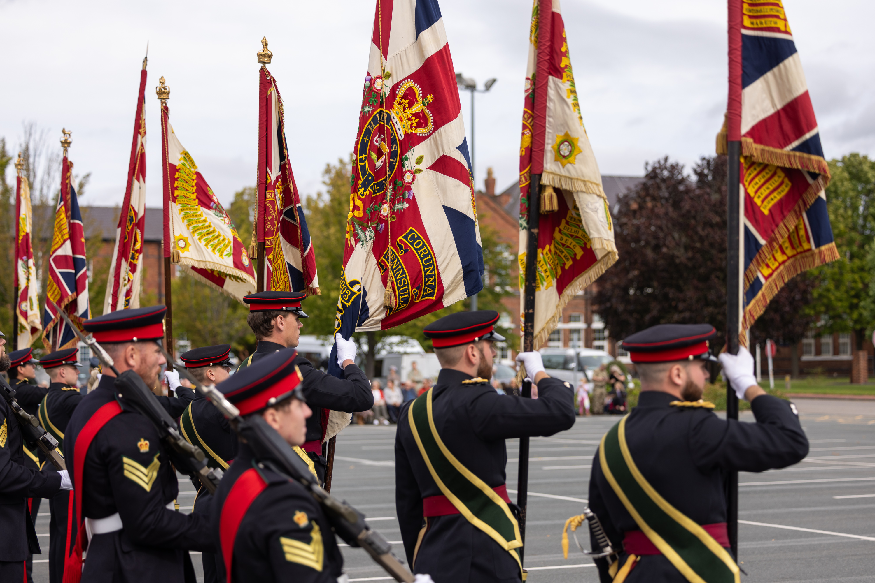 Military personnel in formal dress uniforms march in formation, holding various ceremonial flags. The flags feature intricate designs and patterns, including a prominent Union Jack. The setting appears to be an outdoor event with trees and buildings in the background.