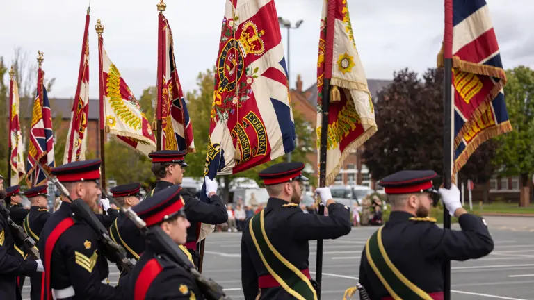 Military personnel in formal dress uniforms march in formation, holding various ceremonial flags. The flags feature intricate designs and patterns, including a prominent Union Jack. The setting appears to be an outdoor event with trees and buildings in the background.