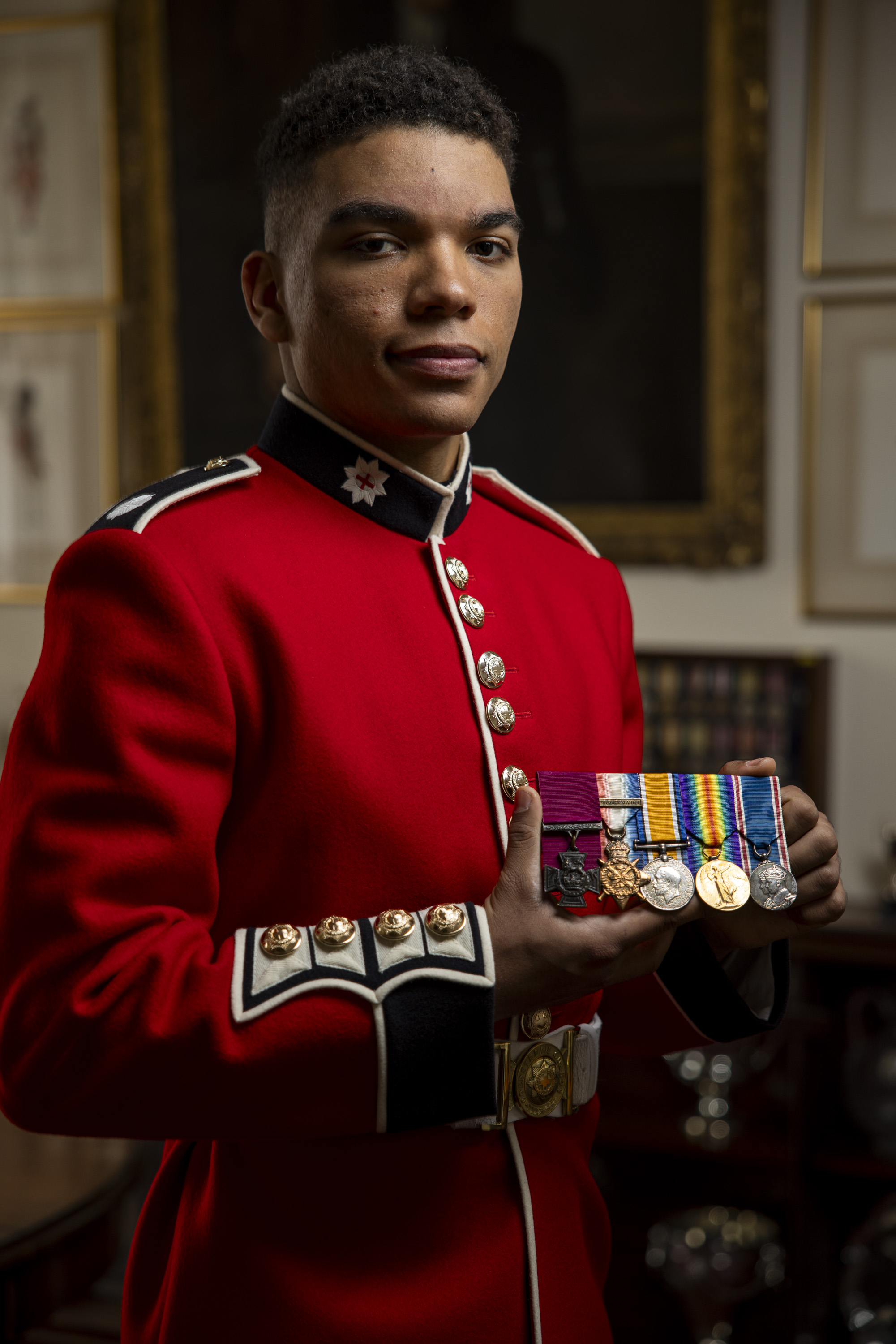 A soldier in a red tunic holds out several medals.
