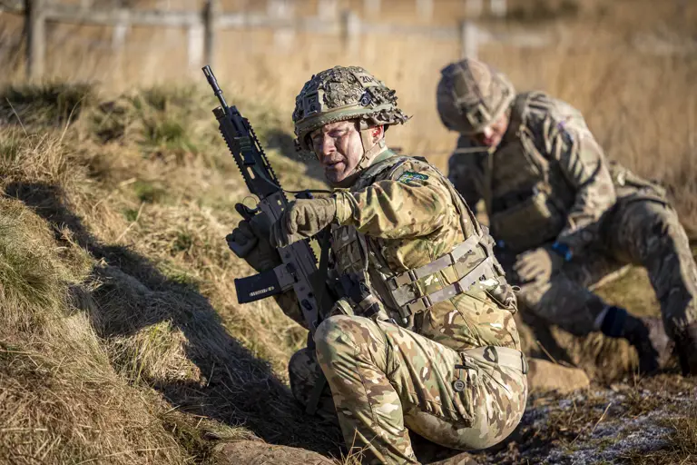 Two soldiers in uniform are pictured during a training exercise, the man closest is holding a gun and pointing.