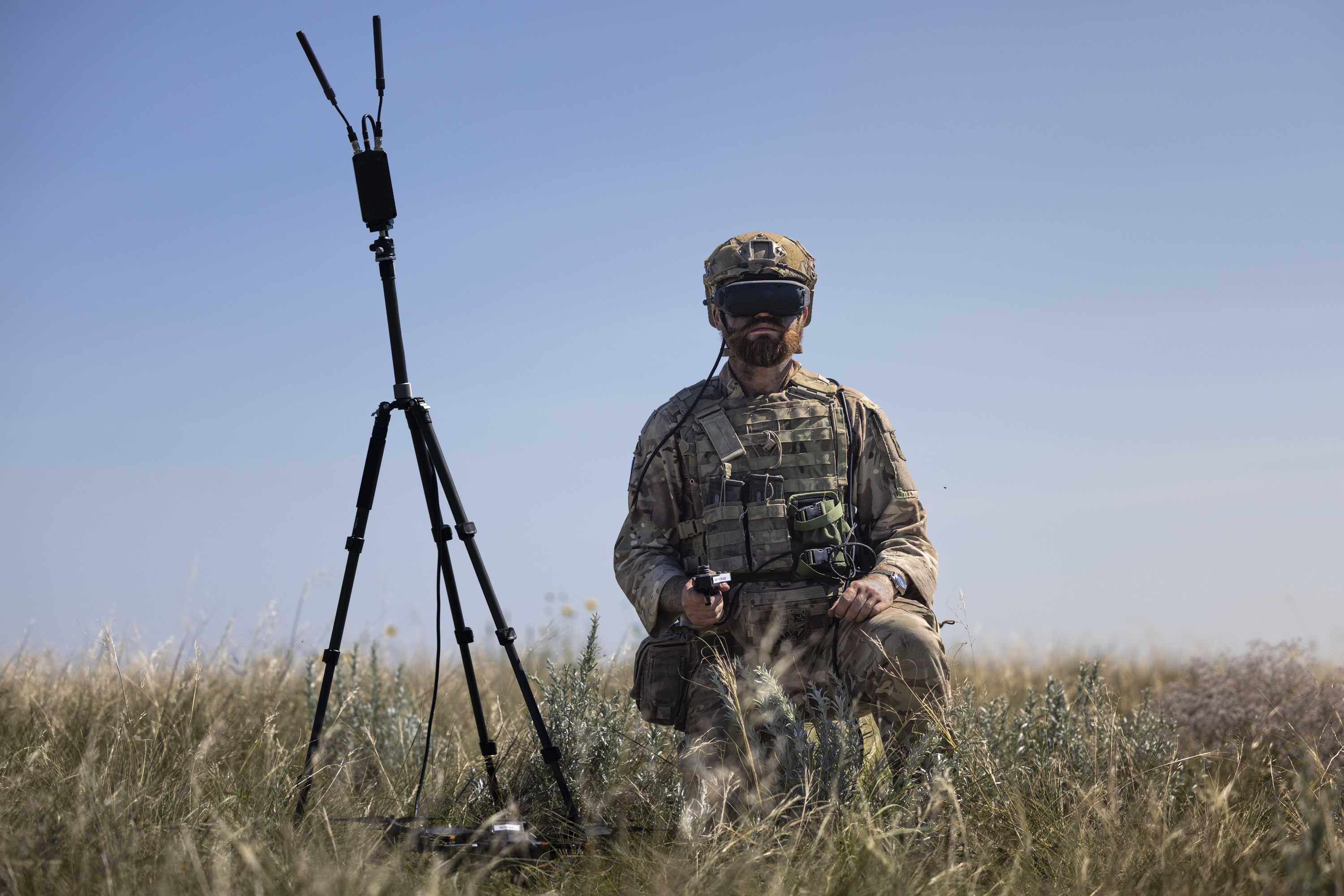 An individual in camouflage gear kneels in a grassy field, holding a controller, with a drone and a tripod-mounted device nearby. The person is wearing a headset with goggles.