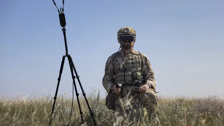 An individual in camouflage gear kneels in a grassy field, holding a controller, with a drone and a tripod-mounted device nearby. The person is wearing a headset with goggles.