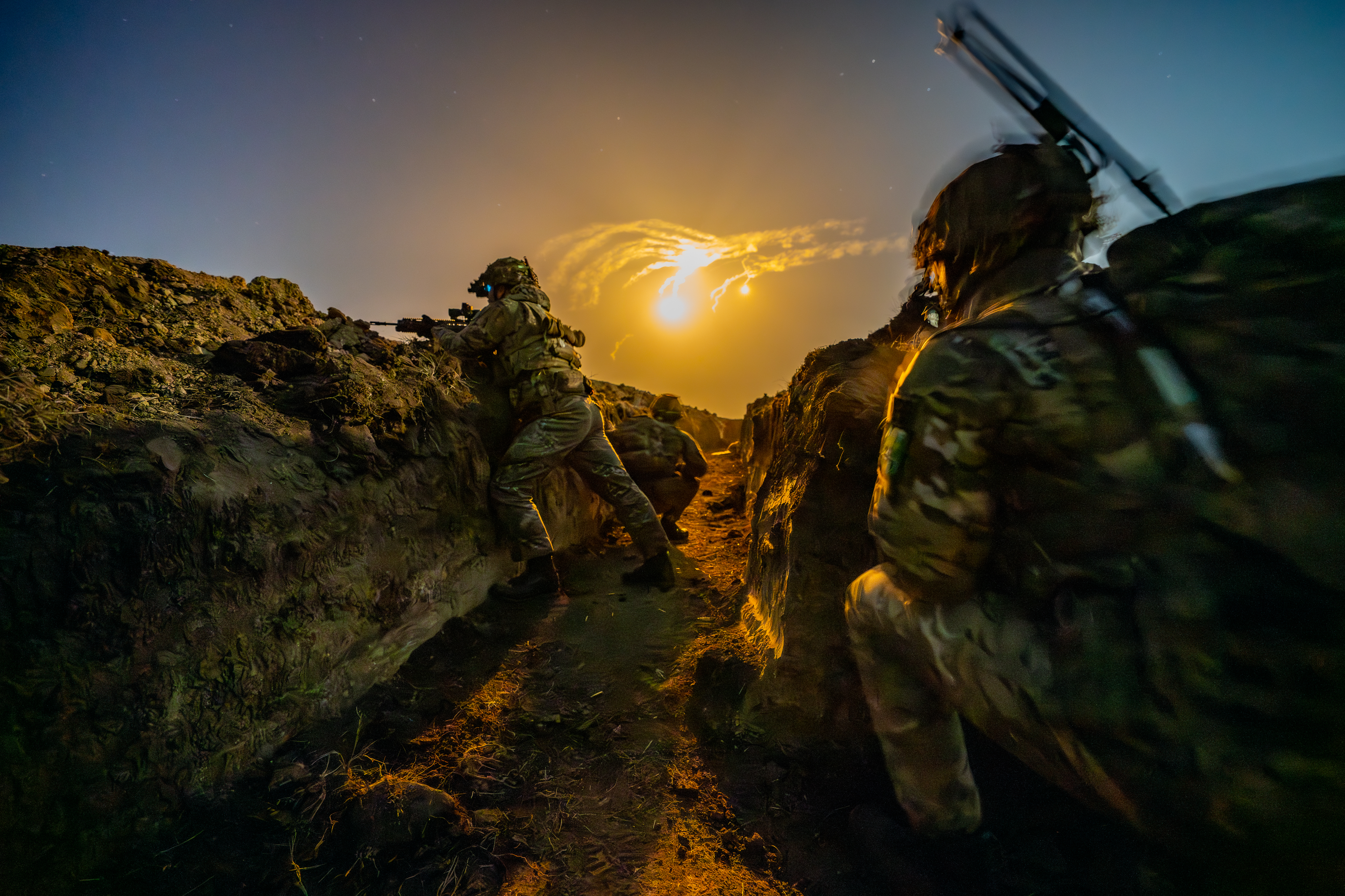 Soldiers carrying out night time live firing training in trenches, they are in full camouflage kit and flares are seen in the night sky.