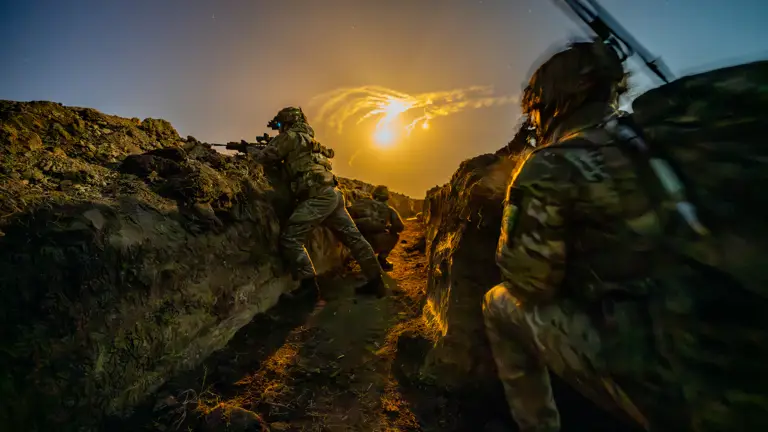 Soldiers carrying out night time live firing training in trenches, they are in full camouflage kit and flares are seen in the night sky.