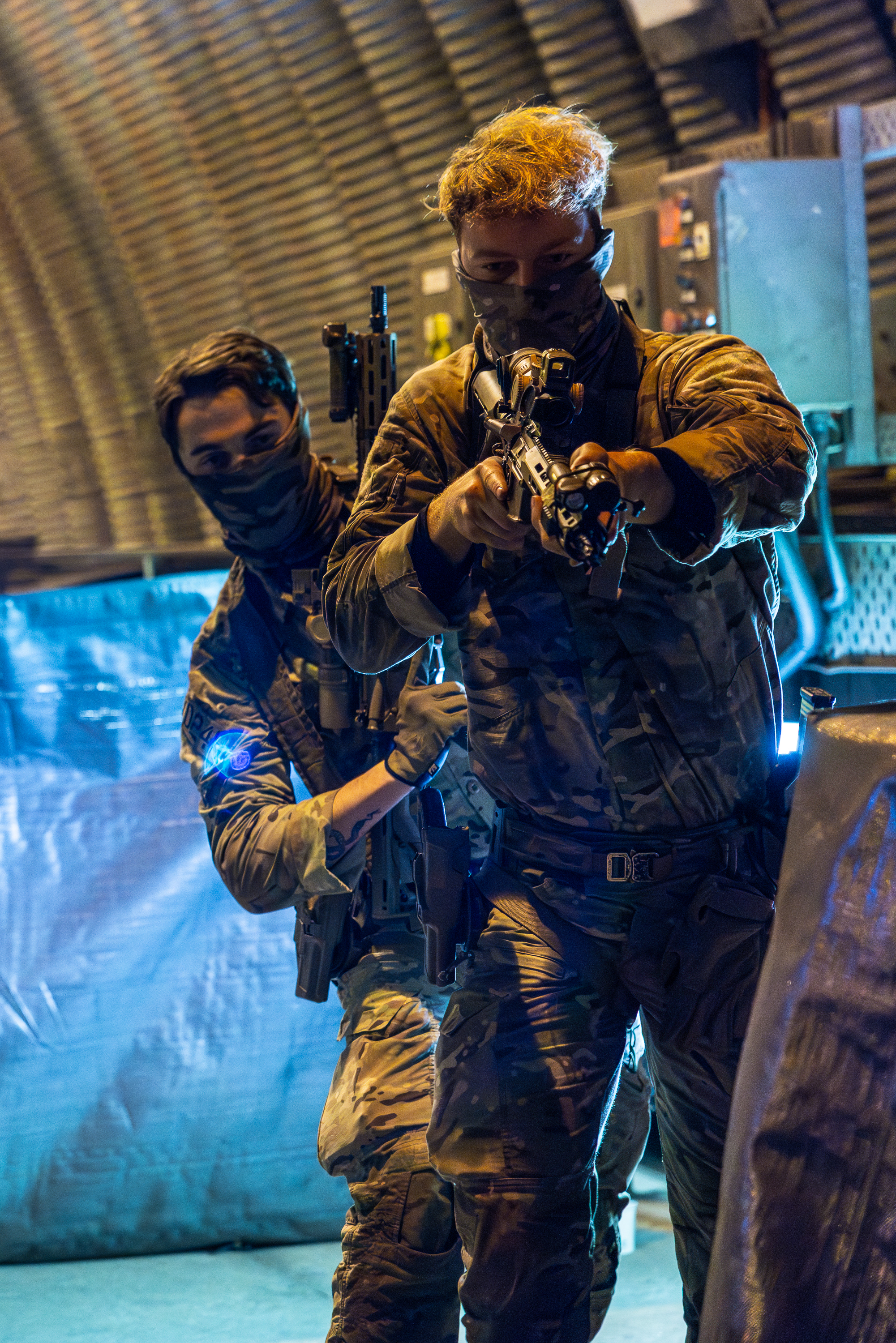Two soldiers in camouflage gear aiming rifles inside a dimly lit military bunker with corrugated metal walls.
