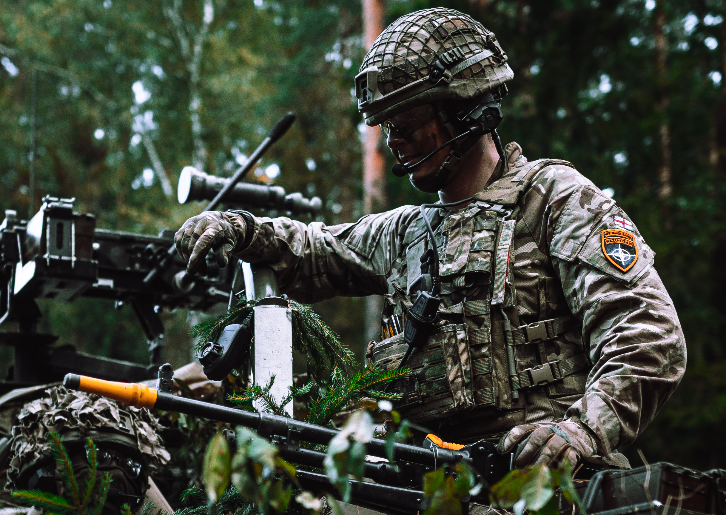 A person in military uniform and helmet is positioned outdoors in a forested area. 