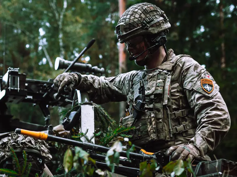A person in military uniform and helmet is positioned outdoors in a forested area.