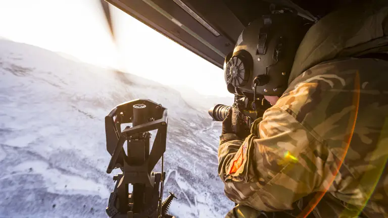 An Army Photographer sits at the door of a flying helicopter filming the snowy landscape beneath him.