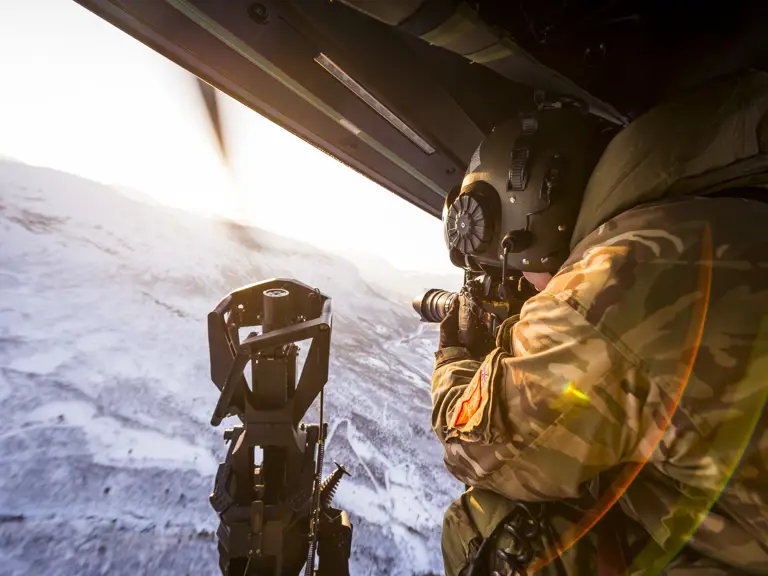 An Army Photographer sits at the door of a flying helicopter filming the snowy landscape beneath him. 