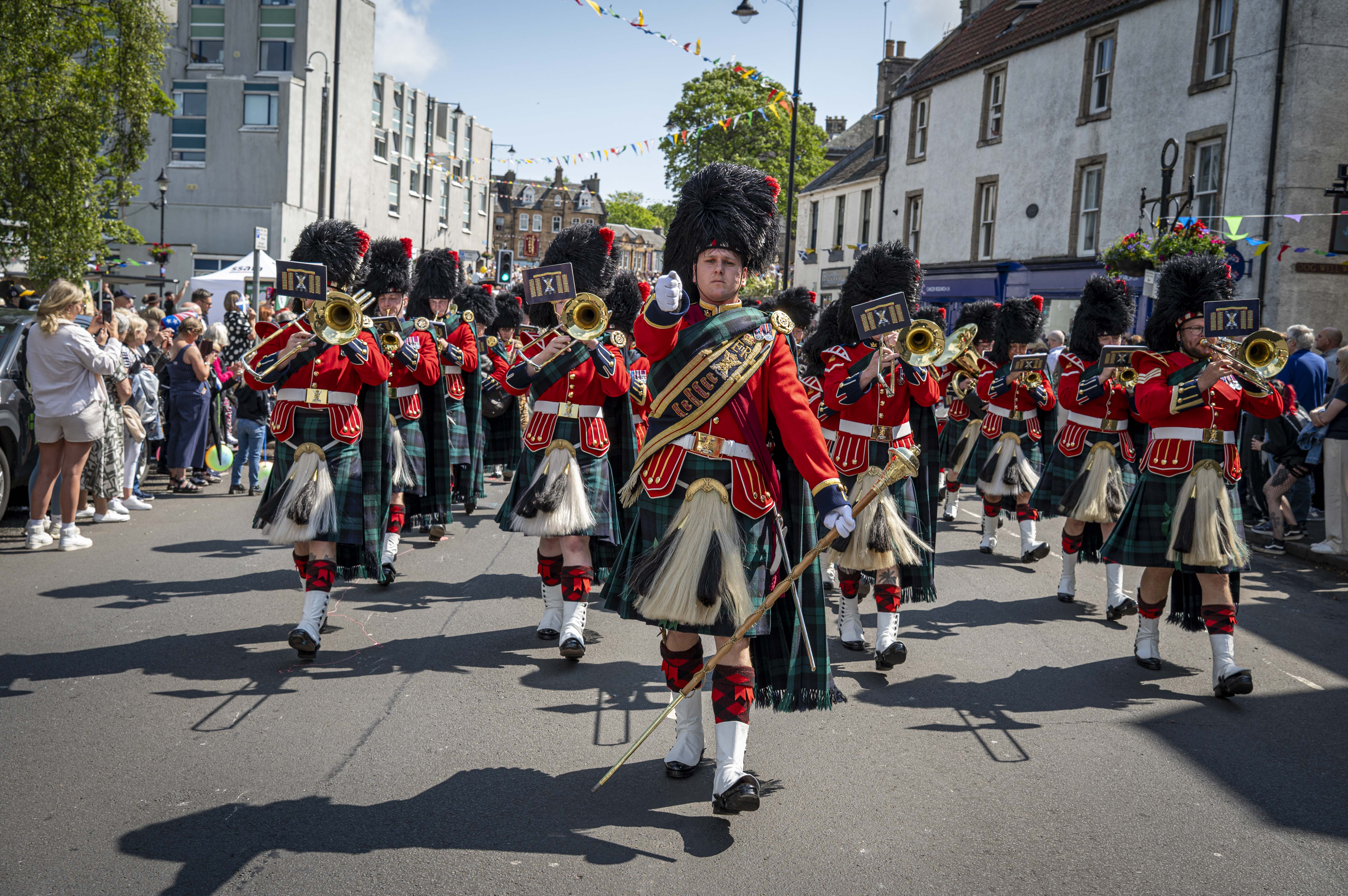 A soldier wears a red ceremonial tunic, sash and a black feather bonnet. He is leading a parade of musicians wearing red tunics and kilts.