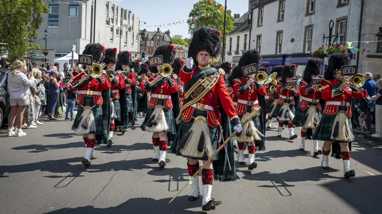 A soldier wears a red ceremonial tunic, sash and a black feather bonnet. He is leading a parade of musicians wearing red tunics and kilts.