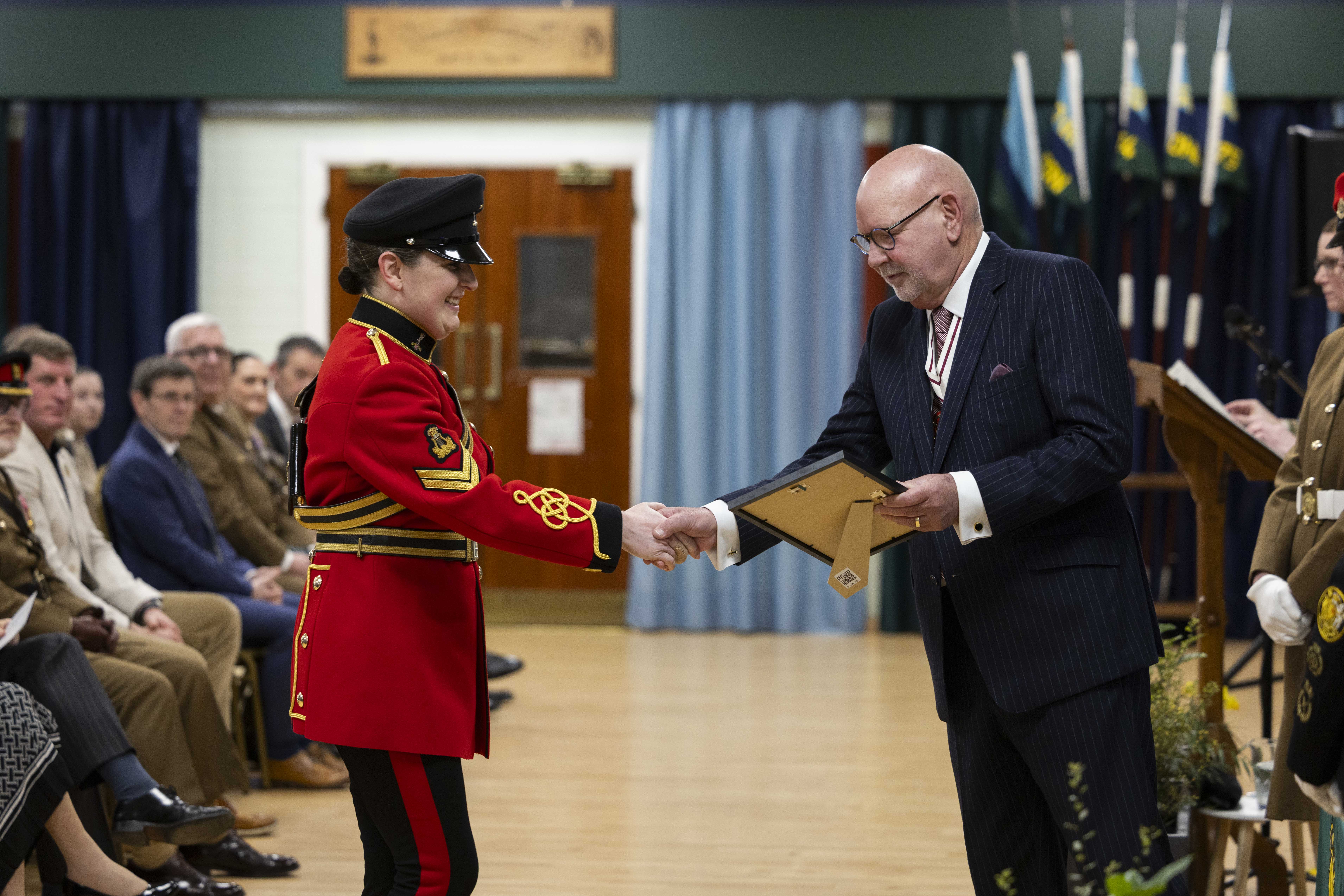 A person in a vibrant red military uniform shakes hands with a bald man in a pinstripe suit, exchanging a certificate at a formal ceremony.