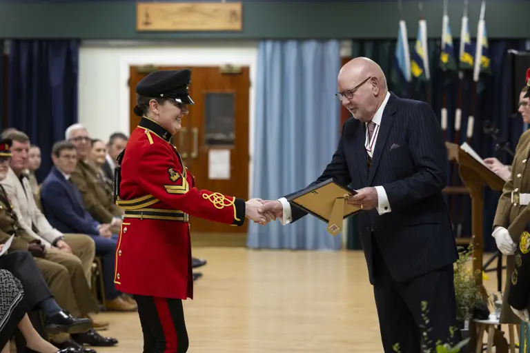 A person in a vibrant red military uniform shakes hands with a bald man in a pinstripe suit, exchanging a certificate at a formal ceremony.