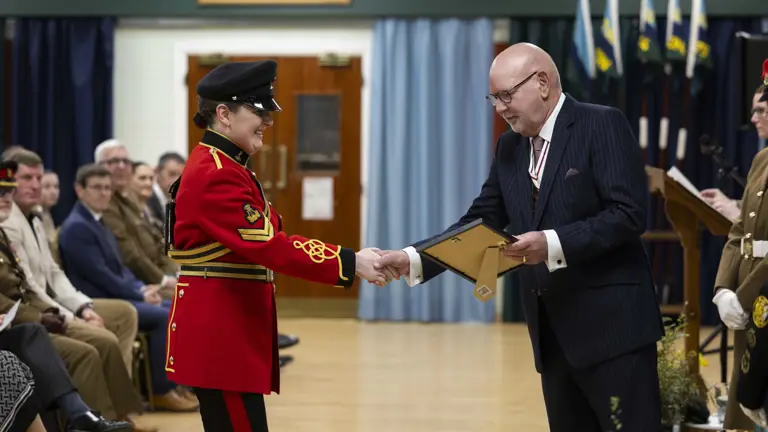 A person in a vibrant red military uniform shakes hands with a bald man in a pinstripe suit, exchanging a certificate at a formal ceremony.