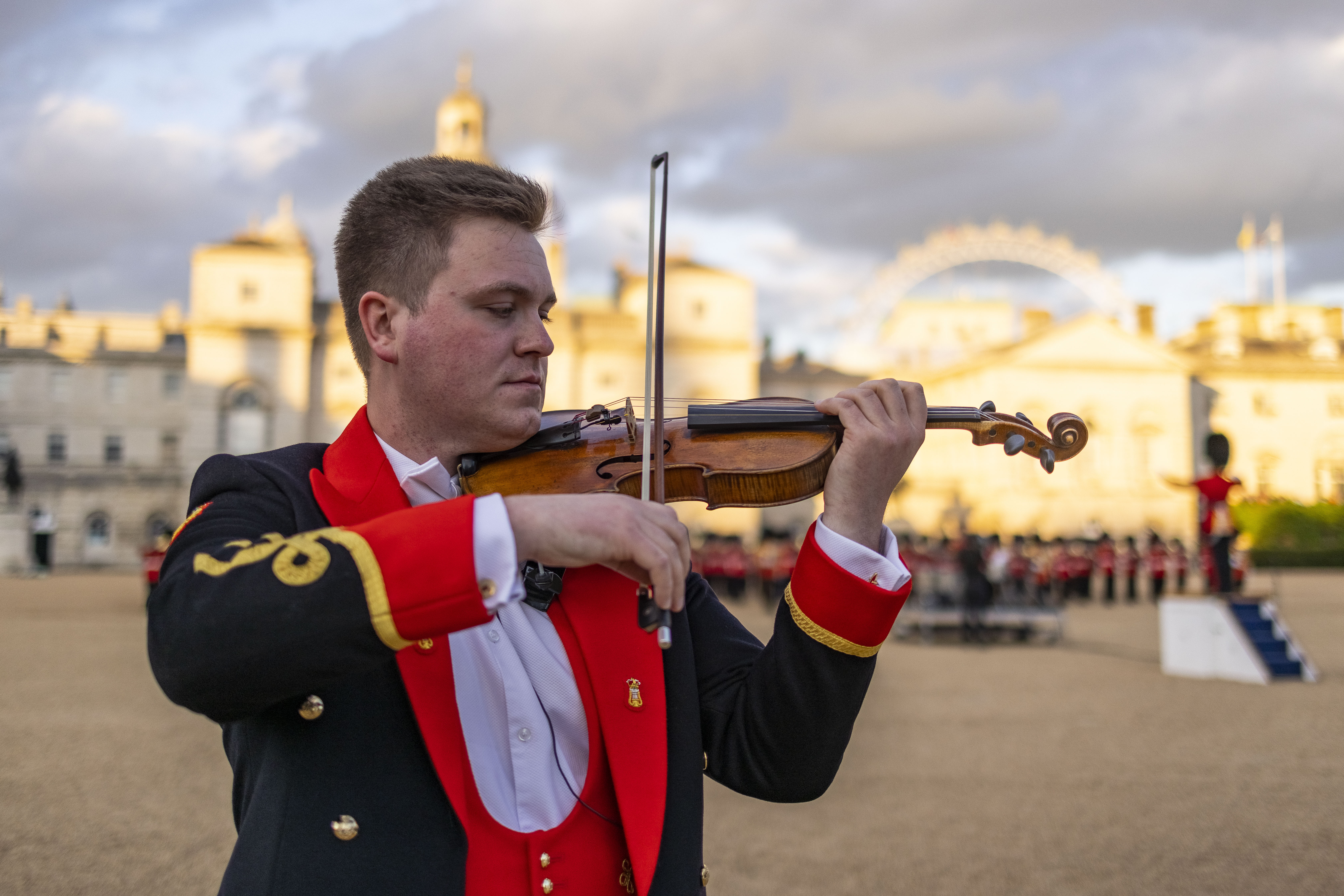 Musician in formal red and black uniform playing violin outdoors with historic buildings and cloudy sky in background.