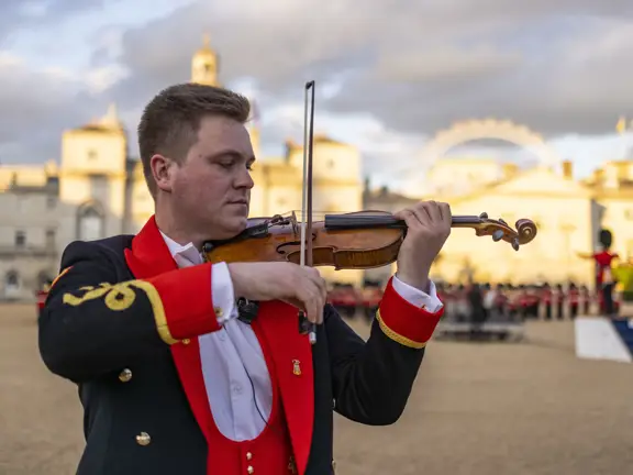 Musician in formal red and black uniform playing violin outdoors with historic buildings and cloudy sky in background.