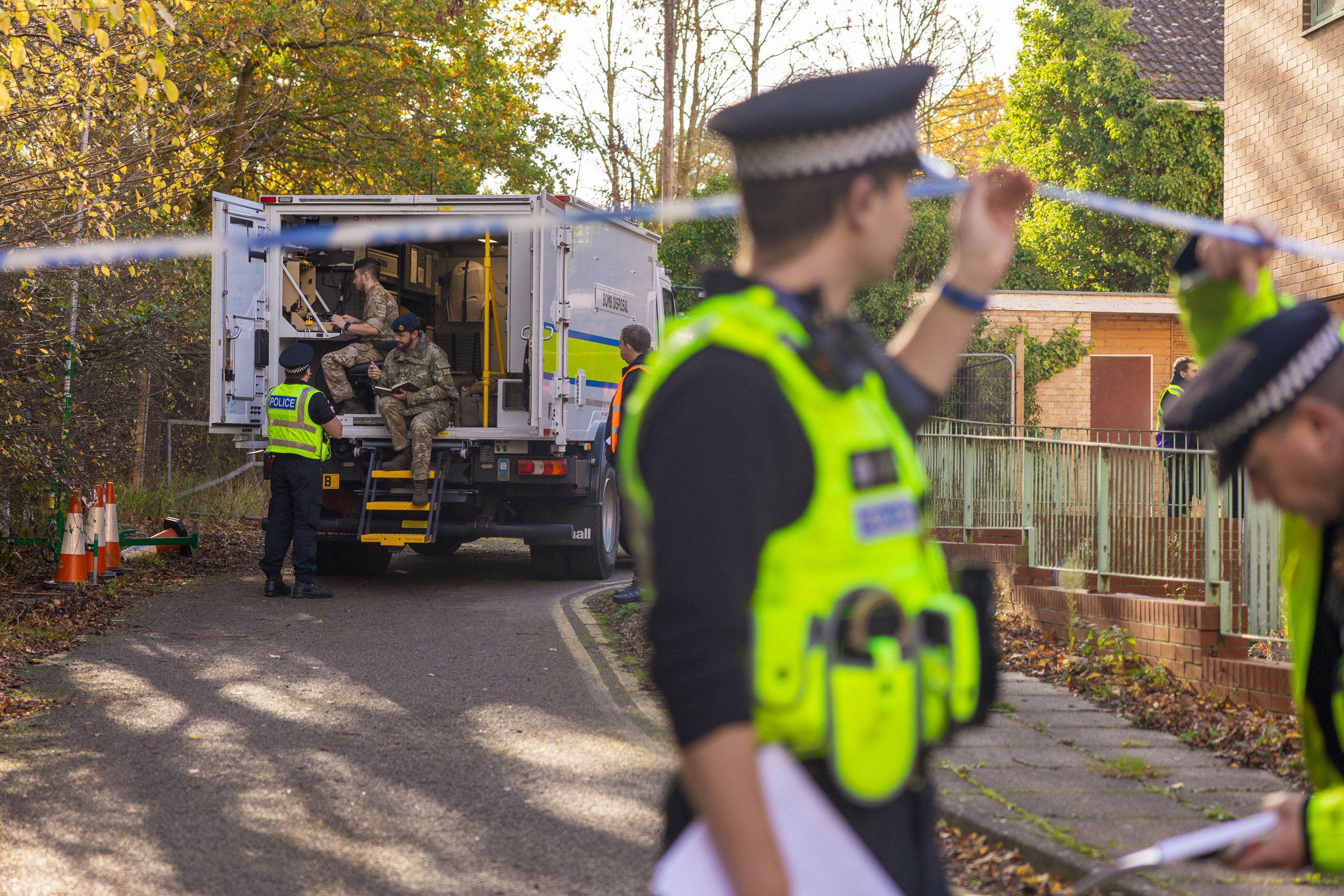 Police officers secure a residential area with caution tape while military personnel unload equipment from a truck.