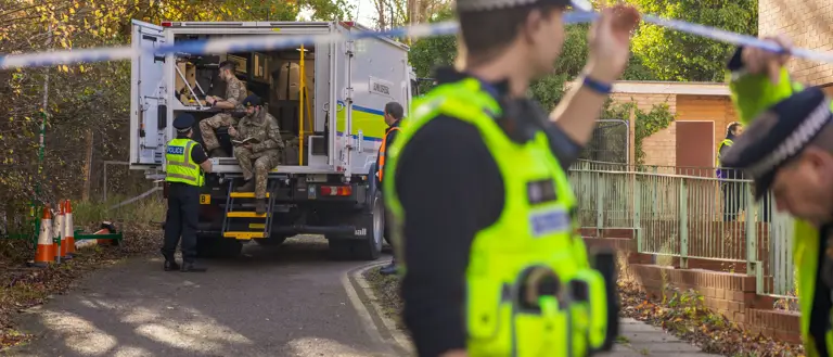 Police officers secure a residential area with caution tape while military personnel unload equipment from a truck.