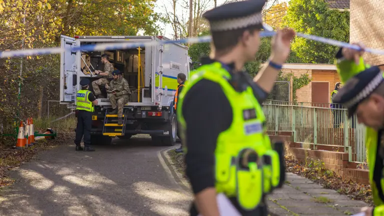 Police officers secure a residential area with caution tape while military personnel unload equipment from a truck.