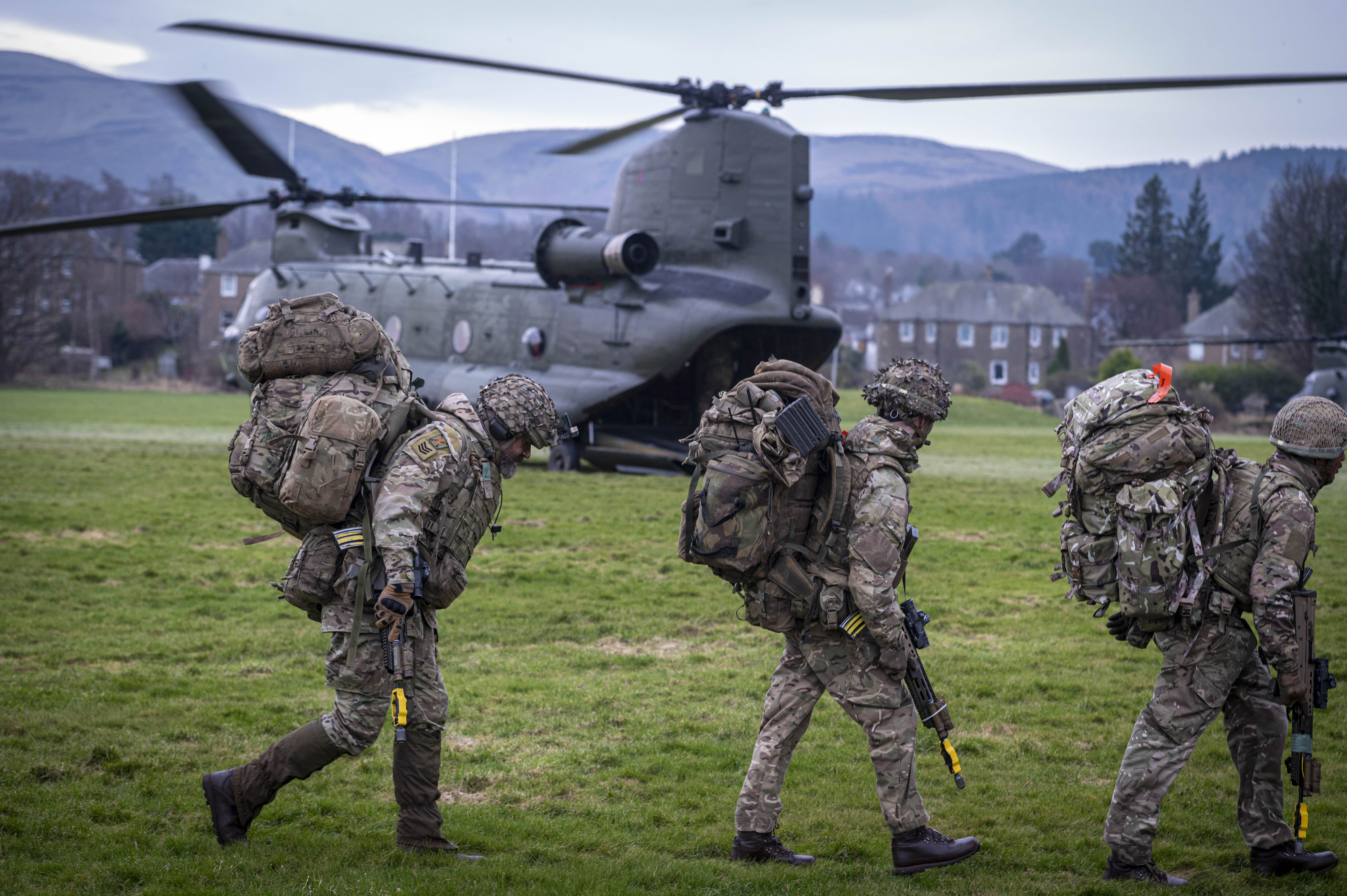 soldiers in camouflage uniform are seen walking in front of a Chinook with rifles and army gear ready to board it in a field.