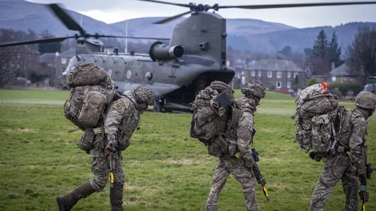 soldiers in camouflage uniform are seen walking in front of a Chinook with rifles and army gear ready to board it in a field.