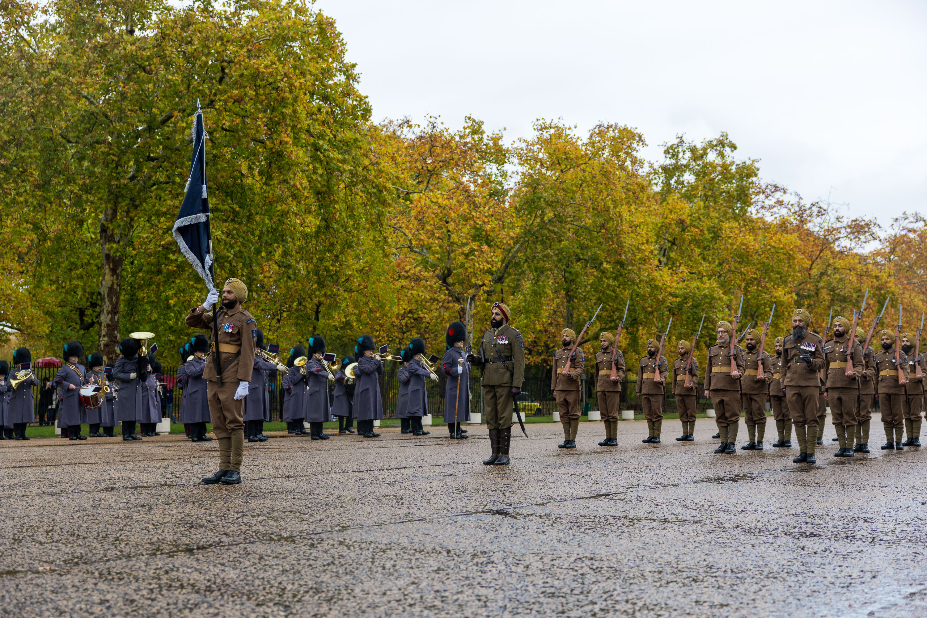 Soldiers in brown uniforms and turbans stand in formation during a ceremonial parade with a band in the background.